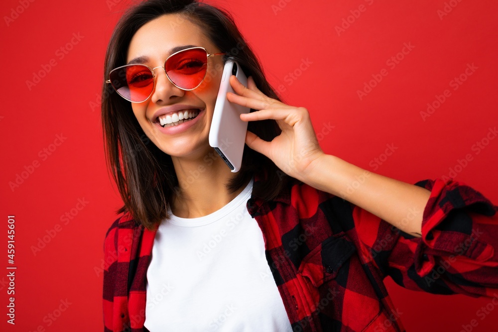 Closeup photo of pretty happy smiling young brunet woman wearing stylish red shirt white t-shirt and red sunglasses isolated over red background communicating on mobile phone looking at camera Closeup photo of pretty happy smiling young brunet woman wearing stylish red shirt white t-shirt and red sunglasses isolated over red background communicating on mobile phone looking at camera