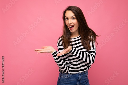 Portrait photo of young happy positive beautiful brunette woman with sincere emotions wearing casual striped pullover isolated on pink background with copy space and pointing hands at free space for © Ivan Traimak