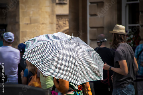 Closeup of woman mostly hidden by bright black and white umbrella walks through crowded street with tourists and performer-singer in background - Selective focus.