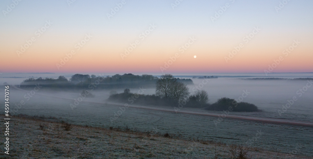 an early morning mist filled valley with the snow moon low in the sunrise sky 