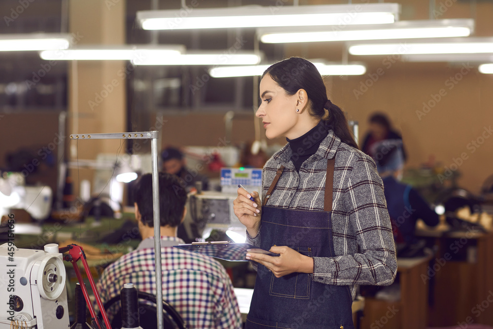 Portrait of a serious young female worker at a shoe making factory. Side view of a sewing workshop manager holding a clipboard and supervising working process at a footwear manufacturing plant