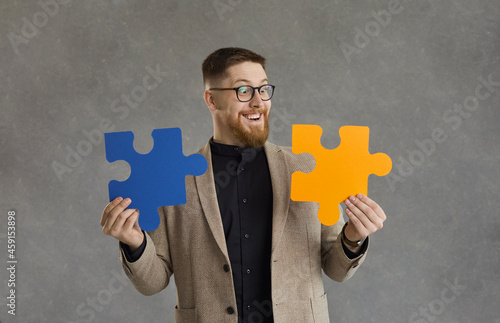 Funny smiling male office worker, makes up two colored puzzle pieces standing on a gray background. Millennial man easily solves difficult problems at work Business solutions and success concept. photo