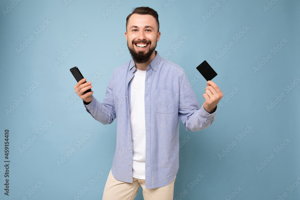Photo shot of positive handsome bearded good looking young man wearing casual stylish outfit poising isolated on background with empty space holding in hand phone and gredit card looking at camera Photo shot of positive handsome bearded good looking young man wearing casual stylish outfit poising isolated on background with empty space holding in hand phone and gredit card looking at camera