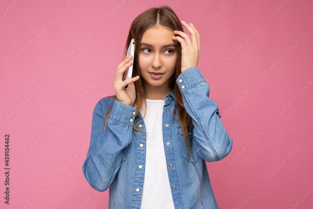 Photo of beautiful sexy thinking young blonde woman wearing casual blue jean shirt isolated over pink background holding in hand and talking on mobile phone looking down to the side Photo of beautiful sexy thinking young blonde woman wearing casual blue jean shirt isolated over pink background holding in hand and talking on mobile phone looking down to the side