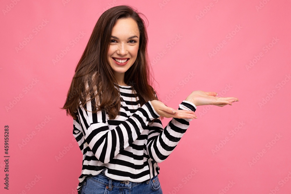 Photo of young happy positive beautiful brunette woman with sincere emotions wearing casual striped pullover isolated on pink background with copy space and pointing at empty space for advertisement Photo of young happy positive beautiful brunette woman with sincere emotions wearing casual striped pullover isolated on pink background with copy space and pointing at empty space for advertisement
