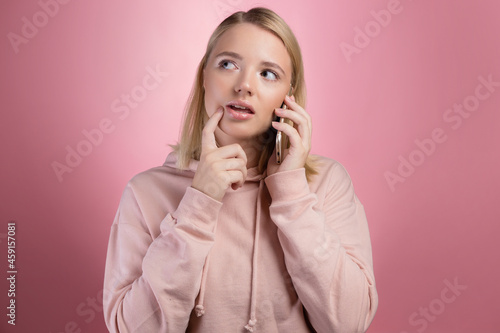 To think about a proposal, a young woman is talking on the phone, thoughtful or doubtful. An attractive young woman in a pink hoodie uses a smartphone, a photo on pink.