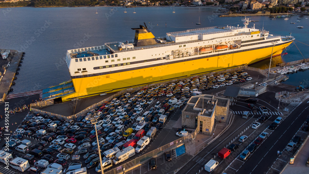 Fototapeta premium Yellow ferry in the port of Île Rousse in Upper Corsica, France - Rows of cars waiting in line to embark and cross the Mediterranean Sea to France or Italy