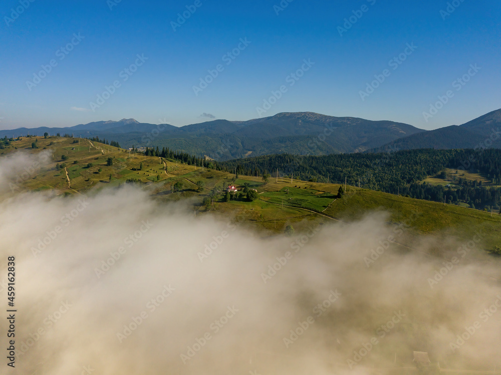 A thin morning fog covers the Ukrainian mountains. Green grass on the slopes of the mountains. A curly thin fog spreads over the mountains. Aerial drone view.