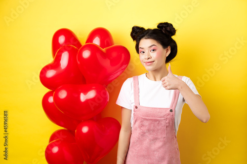 Fashionble asian woman in romantic outfit with make-up, showing thumb up and smiling, praising Valentines day offer, standing near red heart balloons, yellow background photo