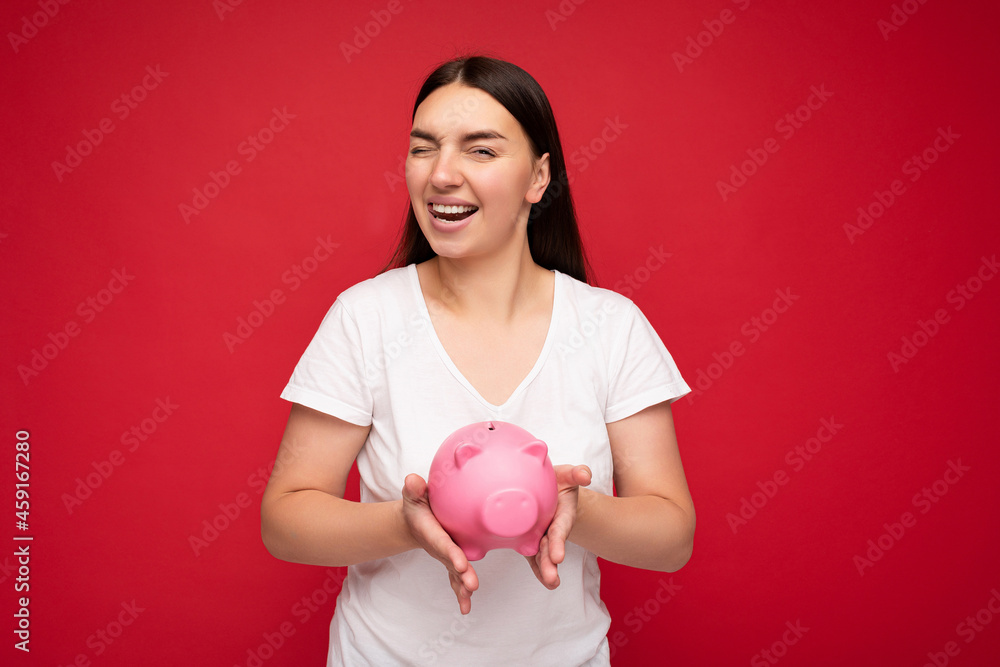 Portrait photo of happy positive smiling young beautiful attractive brunette woman with sincere emotions wearing casual white t-shirt isolated over red background with copy space and holding pink Portrait photo of happy positive smiling young beautiful attractive brunette woman with sincere emotions wearing casual white t-shirt isolated over red background with copy space and holding pink