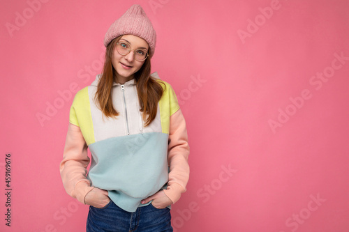Photo of pbeautiful positive happy young dark blonde female person isolated over pink background wall wearing stylish hoodie pink hat and optical glasses looking at camera