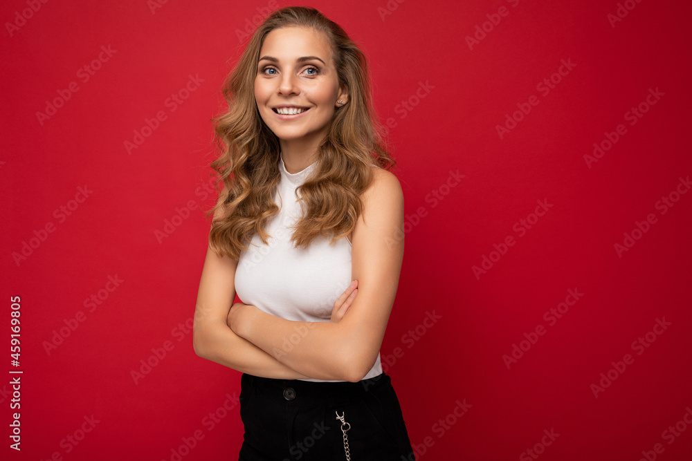 Portrait of young beautiful pretty happy joyful smiling blonde woman with sincere emotions wearing casual white top isolated over red background with empty space with arms crossed