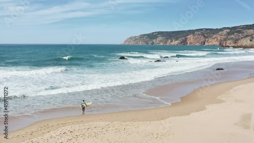 Scenic view of big ocean waves washing mountain area. Overhead shot of male athlete holding surfboard in hands. Young surfer entering sea for training on sunny day, 4k footage with copy space