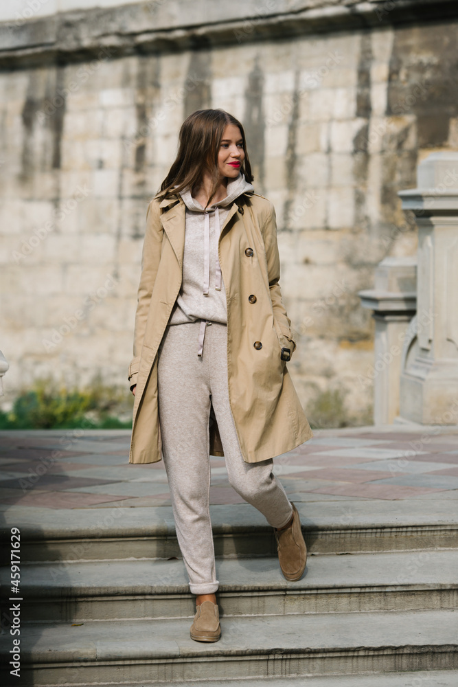 Fototapeta premium Portrait of fashionable women in beige sports suit, trench coat and stylish suede loafer posing on the stone stairs