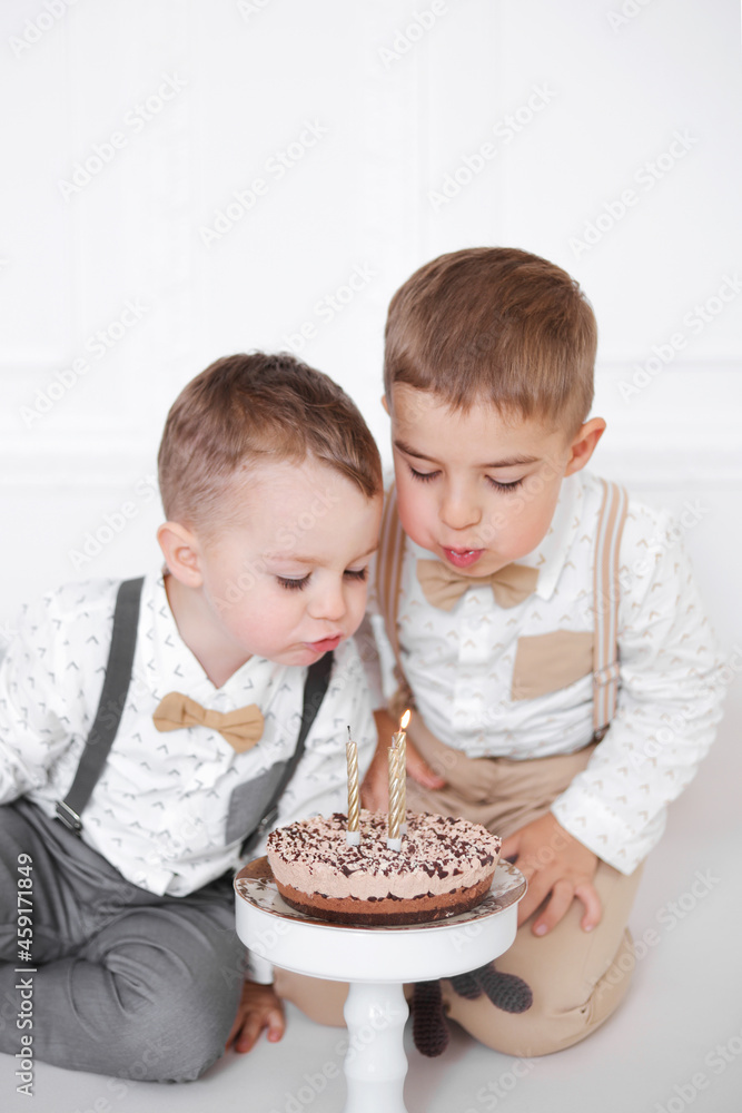 Two boys celebrating birthday, children have a B-day party. Kids blowing candles on the birthday cake. Celebration, white minimalist interior. Two boys celebrating birthday, children have a B-day party. Kids blowing candles on the birthday cake. Celebration, white minimalist interior.