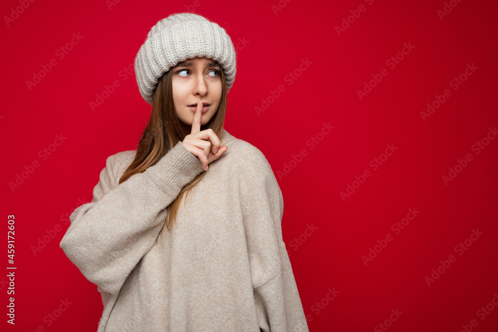 Portrait of positive young beautiful dark blonde woman with sincere emotions wearing beige pullover and knitted hat isolated over red background with copy space and asking remain silent with shhh