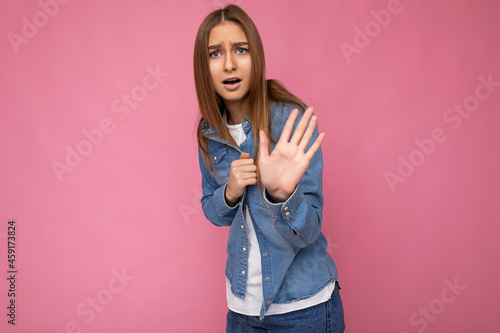 Portrait of dissatisfied shocked amazed young attractive dark blond woman with sincere emotions wearing blue denim shirt isolated over pink background with copy space and hiding showing stop sigh with
