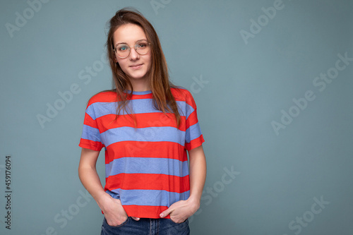 Photo shot of beautiful young brunette woman wearing optical glasses casual srtiped pink and blue t-shirt standing isolated over blue background wall and looking at camera photo