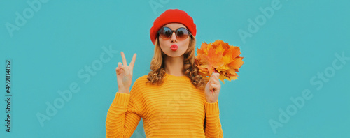 Autumn portrait of beautiful young woman with yellow maple leaves blowing her lips wearing a knitted sweater, red beret on blue background