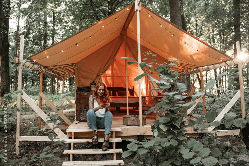 The girl sits next to the tent and chatting messaging streaming on the smartphone, posting, liking in social media. Travel outside city in the woods. Camping. Low-budget vacation in glamping tent The girl sits next to the tent and chatting messaging streaming on the smartphone, posting, liking in social media. Travel outside city in the woods. Camping. Low-budget vacation in glamping tent
