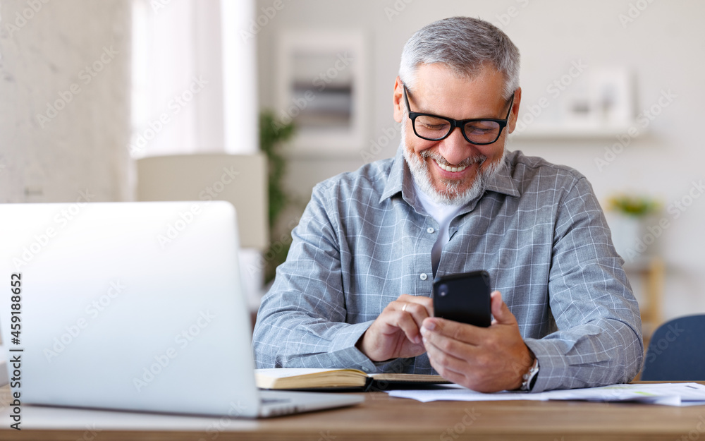 Smiling senior man using mobile phone while sitting at his cozy workplace with laptop at home Smiling senior man using mobile phone while sitting at his cozy workplace with laptop at home