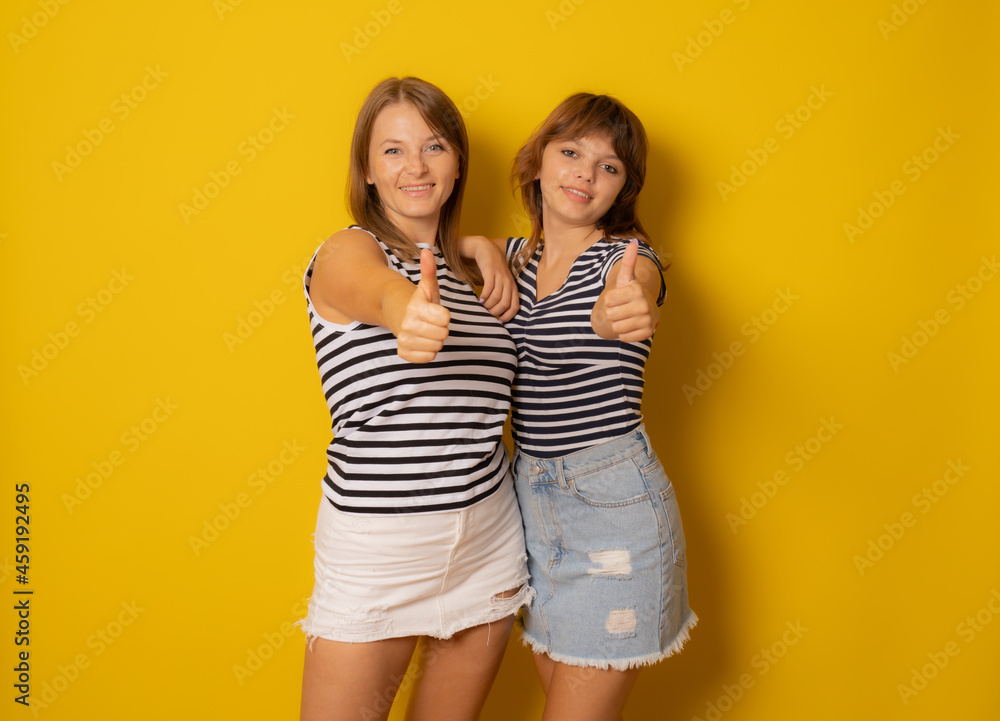 Young mother and her daughter in striped shirts with thumbs up over yellow background. Young mother and her daughter in striped shirts with thumbs up over yellow background.