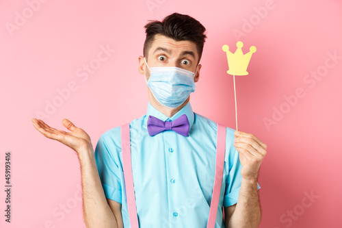 Covid, pandemic and quarantine concept. Clueless and confused young man in medical mask shrugging, showing little party crown, know nothing, standing on pink background photo