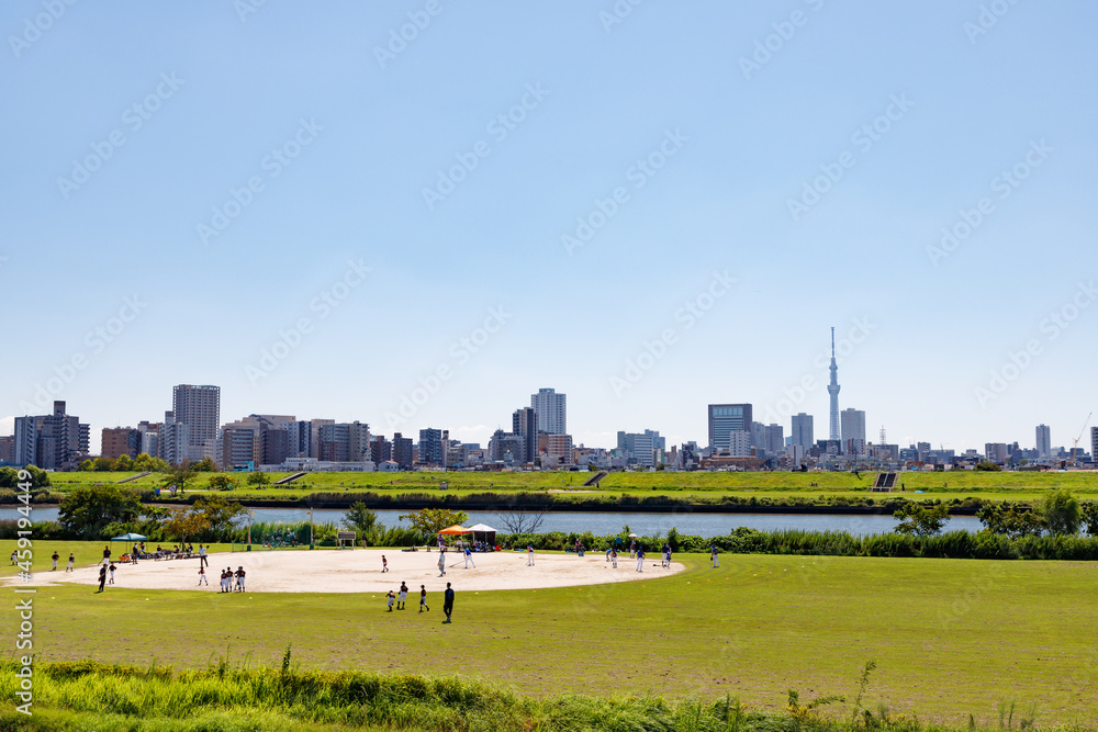 青空を背景に足立区側から見た荒川区方面の景色と河原のグランドで野球をする人々