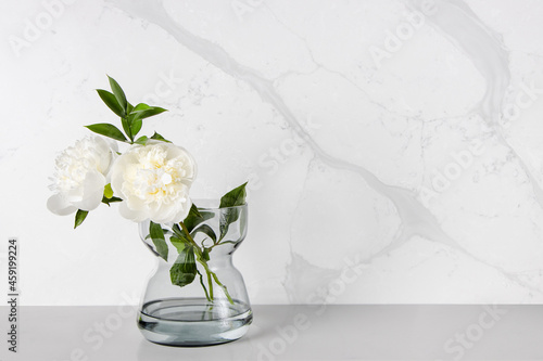 A couple of white peony flowers being placed in a transparent glass vase against a marble-veining kitchen backsplash which was made of quartz stones
