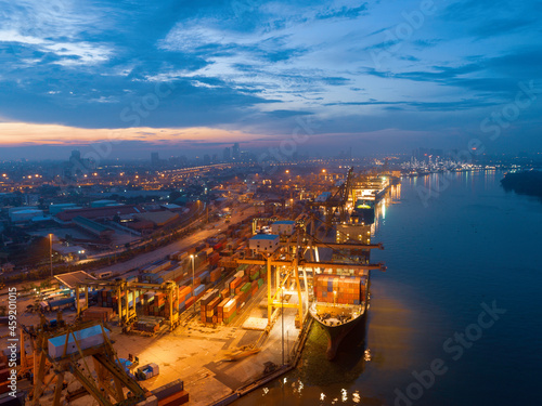 Container cargo ship with ports crane bridge in harbor and refinery industrial at twilight sunset sky logistics and transportation concept