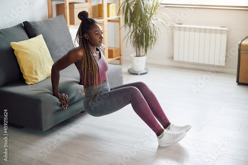 African American sportswoman does triceps dips while leaning on sofa at home. photo
