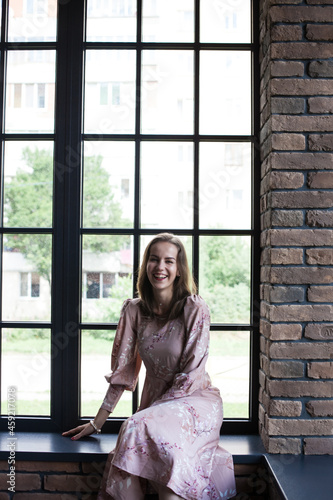 Beautiful young woman in pink dress resting on the windowsill . Happy smiling girl sits on the stained glass window at industrial loft wooden brick style interior. Leisure concept.