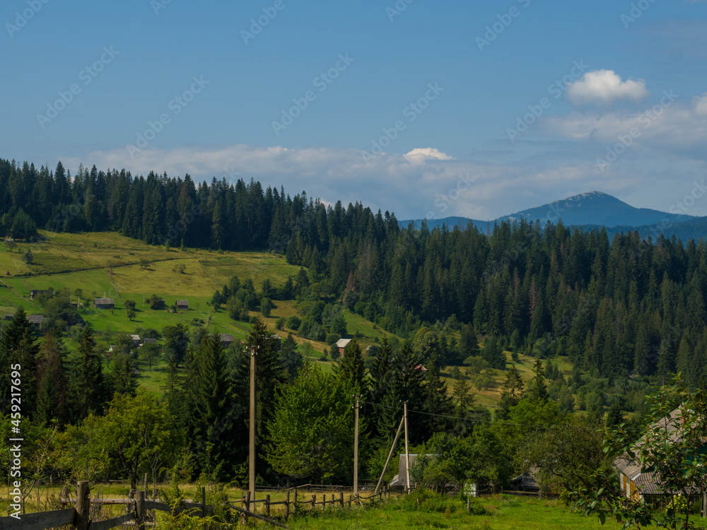 Fototapeta premium Carpathian landscape with cloudy sky. A wooden house on a green meadow in mountains near old forest. Lifestyle in the Carpathian village. Ecology protection concept. Explore the beauty of the world.