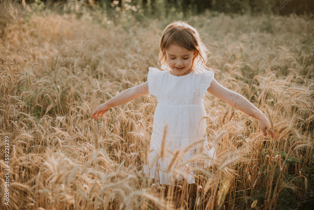 Adorable little blonde girl in a hat and a white linen dress, standing on a wheat field and touching the ears of wheat with her hands. Baby outdoor enjoying nature wheat field. High quality photo Adorable little blonde girl in a hat and a white linen dress, standing on a wheat field and touching the ears of wheat with her hands. Baby outdoor enjoying nature wheat field. High quality photo