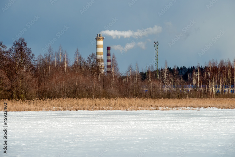 Smoke from chimneys at a factory over the forest near a frozen lake in winter.
