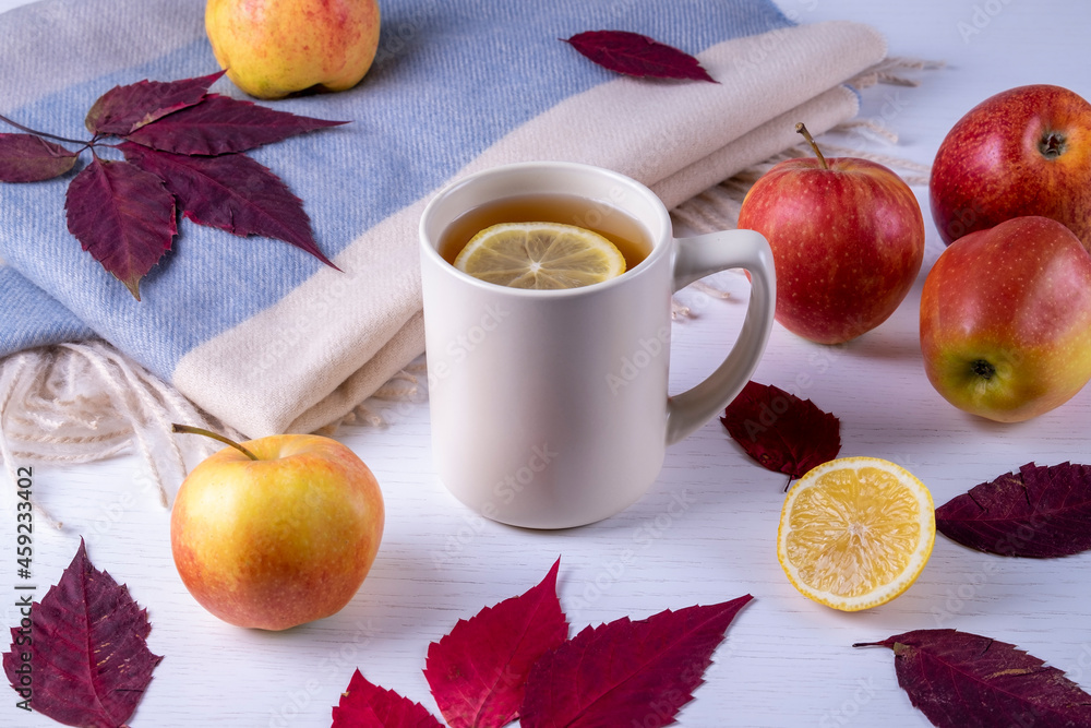 Autumn composition of ceramic mug with hot tea with lemon, soft warm scarf in pastel colors, dry autumn leaves and ripe apples. Selective focus. Autumn composition of ceramic mug with hot tea with lemon, soft warm scarf in pastel colors, dry autumn leaves and ripe apples. Selective focus.