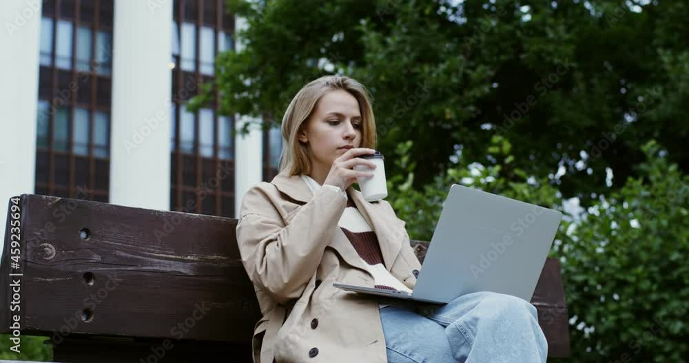 Young woman works at a laptop, sitting on a park bench and drinking coffee Young woman works at a laptop, sitting on a park bench and drinking coffee