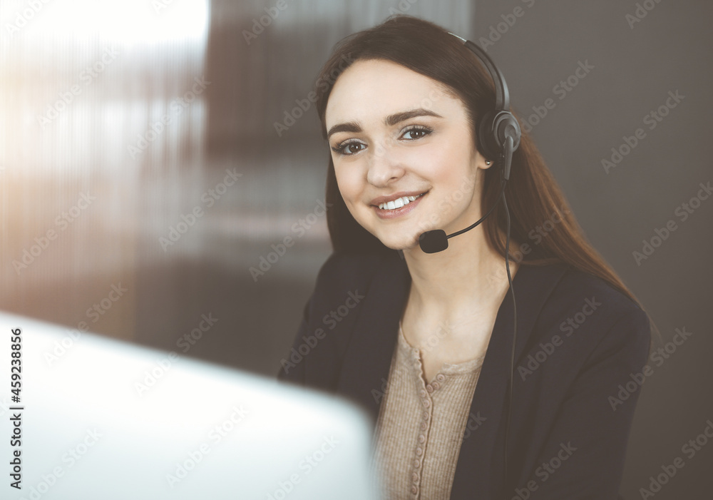 Young friendly girl in headsets is talking to a firm's client, while sitting at the desk in sunny office. Call center operators at work Young friendly girl in headsets is talking to a firm's client, while sitting at the desk in sunny office. Call center operators at work