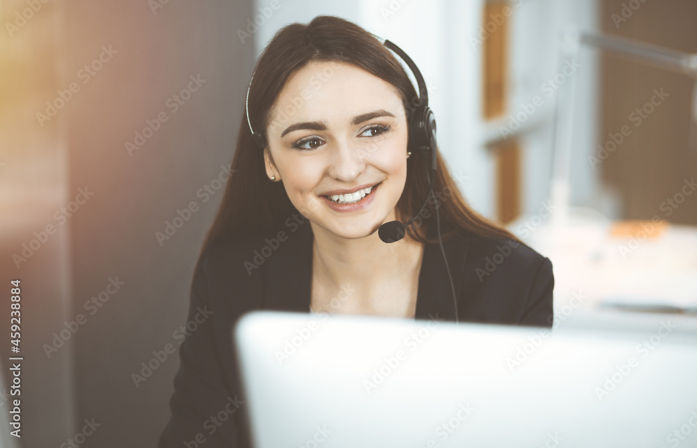 Young friendly girl in headsets is talking to a firm's client, while sitting at the desk in sunny office. Call center operators at work Young friendly girl in headsets is talking to a firm's client, while sitting at the desk in sunny office. Call center operators at work