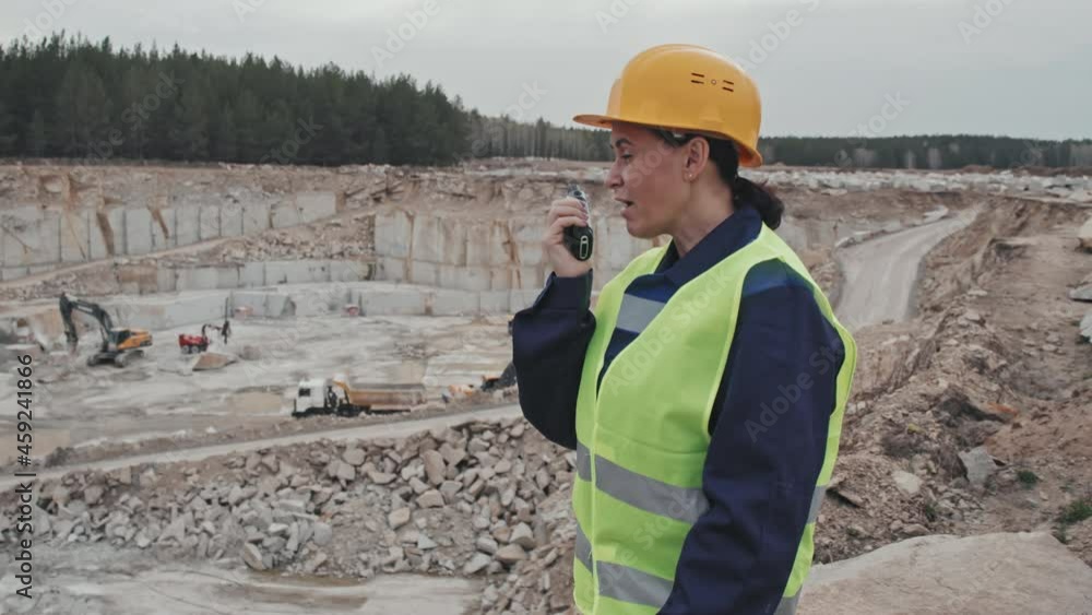 Slowmo tracking of female supervisor in safety vest and hard hat standing on top of granite quarry and using walkie-talkie Slowmo tracking of female supervisor in safety vest and hard hat standing on top of granite quarry and using walkie-talkie