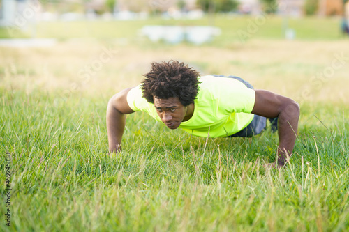 Black man doing push-ups exercising his chest on the grass of an urban park.