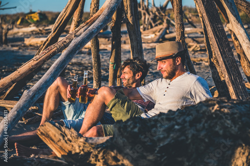 Two young friends spending time together sitting on the beach, drinking beer and toasting on vacation in twilight summer sunset. Friendship concept