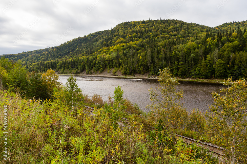 Railway track running along the Matapedia River at the foot of Chic-Chocs mountains covered in mixed trees seen during the early fall cloudy day, Quebec, Canada