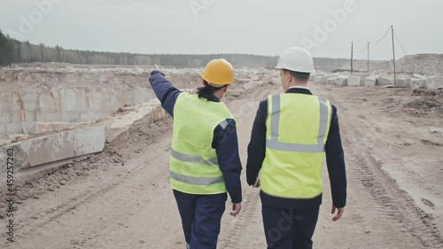 Slowmo tracking of female supervisor and male manager in safety vests and hard hats walking past granite quarry and discussing mining progress