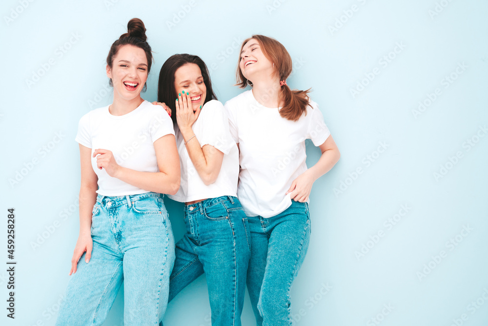 Three young beautiful smiling hipster female in trendy same summer white t-shirt and jeans clothes. Sexy carefree women posing near light blue wall in studio. Cheerful and positive models having fun Three young beautiful smiling hipster female in trendy same summer white t-shirt and jeans clothes. Sexy carefree women posing near light blue wall in studio. Cheerful and positive models having fun