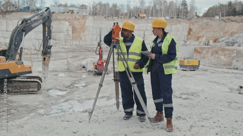 Slowmo tracking of female supervisor making notes on tablet and talking male colleague using surveying measuring equipment in granite quarry. Drill rigs in background Slowmo tracking of female supervisor making notes on tablet and talking male colleague using surveying measuring equipment in granite quarry. Drill rigs in background