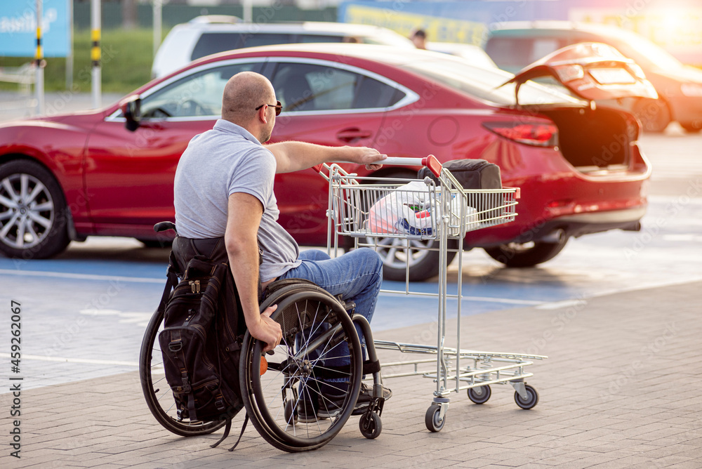Fototapeta premium Adult disabled man in a wheelchair pushes a cart towards a car in a supermarket parking lot