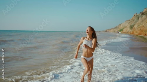happy carefree slender woman with beautiful fitbody running along seashore of sea with waves after storm, girl jogging and jumping through waves photo