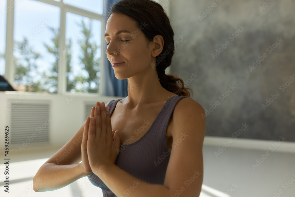 Close-up portrait of woman with beautiful face and clear skin half-turned to camera meditating holding hands in praying position, sitting against background with wall and window of sport studio