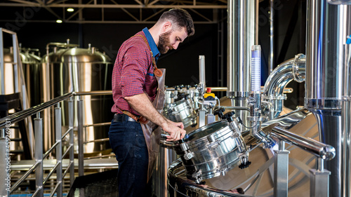 Young male brewer in leather apron supervising the process of beer fermentation at modern brewery factory